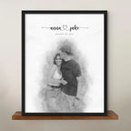 Sketch-style portrait of a couple, the man kissing the woman's forehead, in a black frame on a wooden shelf