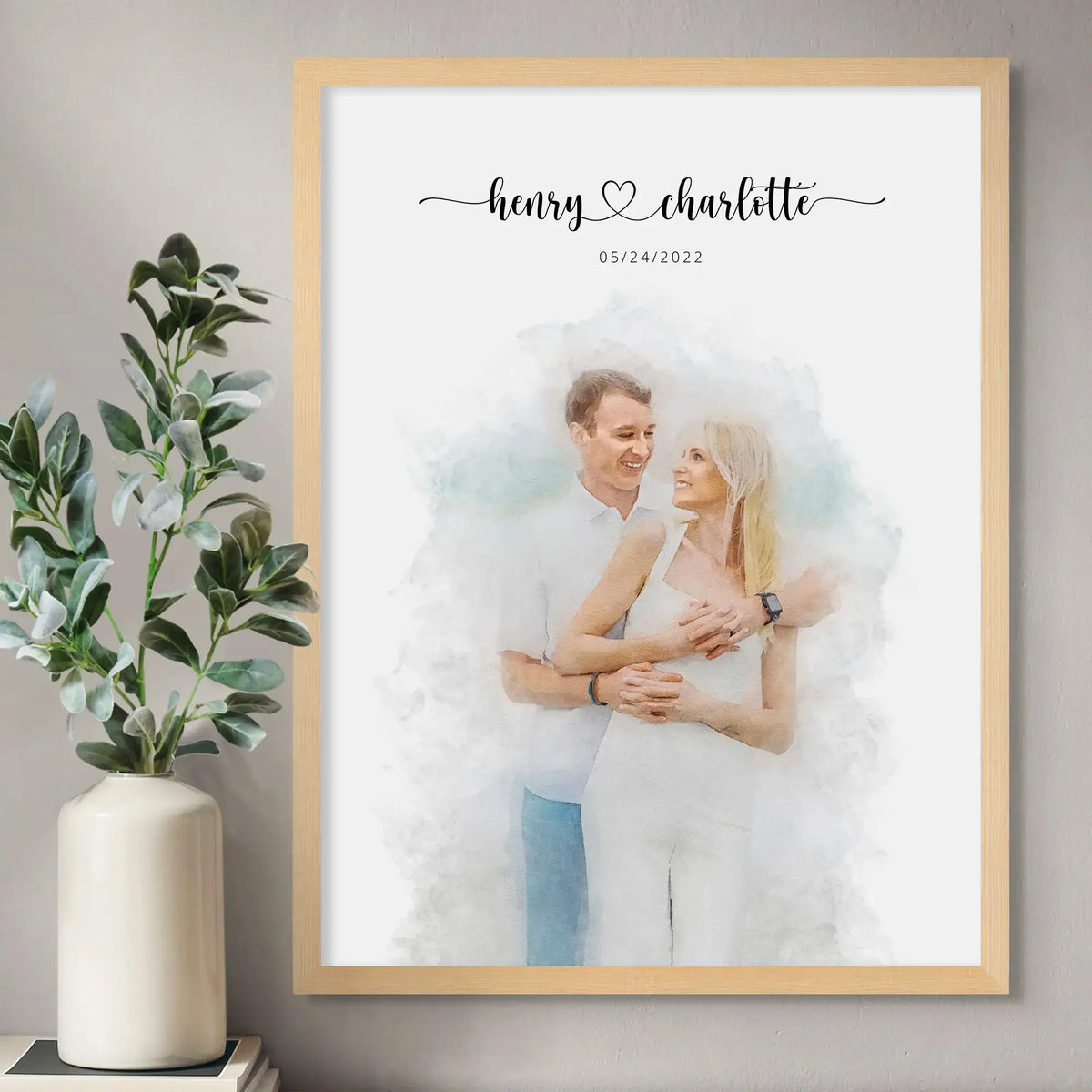 Watercolor-style portrait of a smiling couple dressed in white, in a light wood frame beside a vase of leafy stems