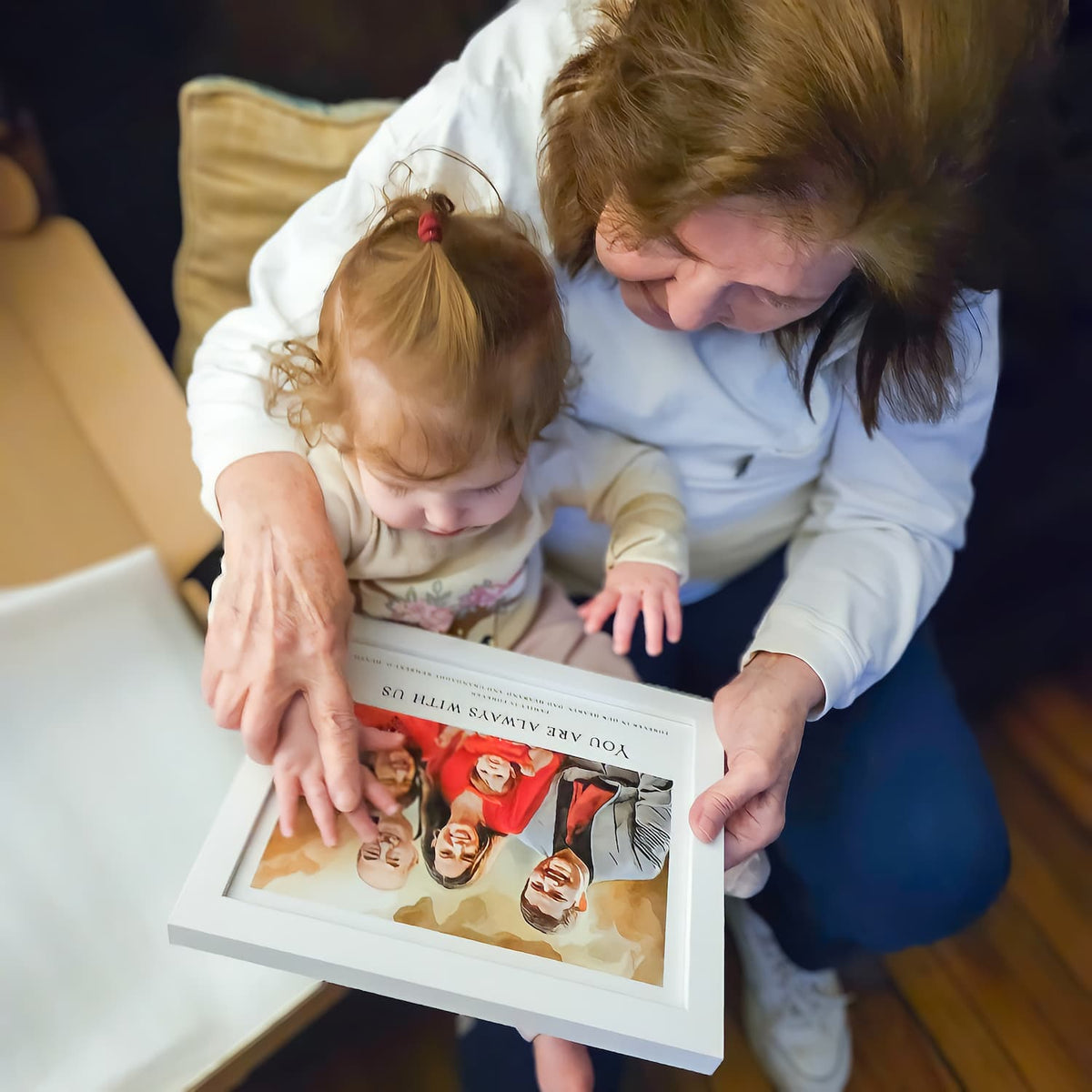 A woman and a toddler look at a white-framed watercolor-style family portrait on her lap titled 'You Are Always With Us'